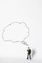 A miniature businessman stands pondering under a large thought bubble on a white background.